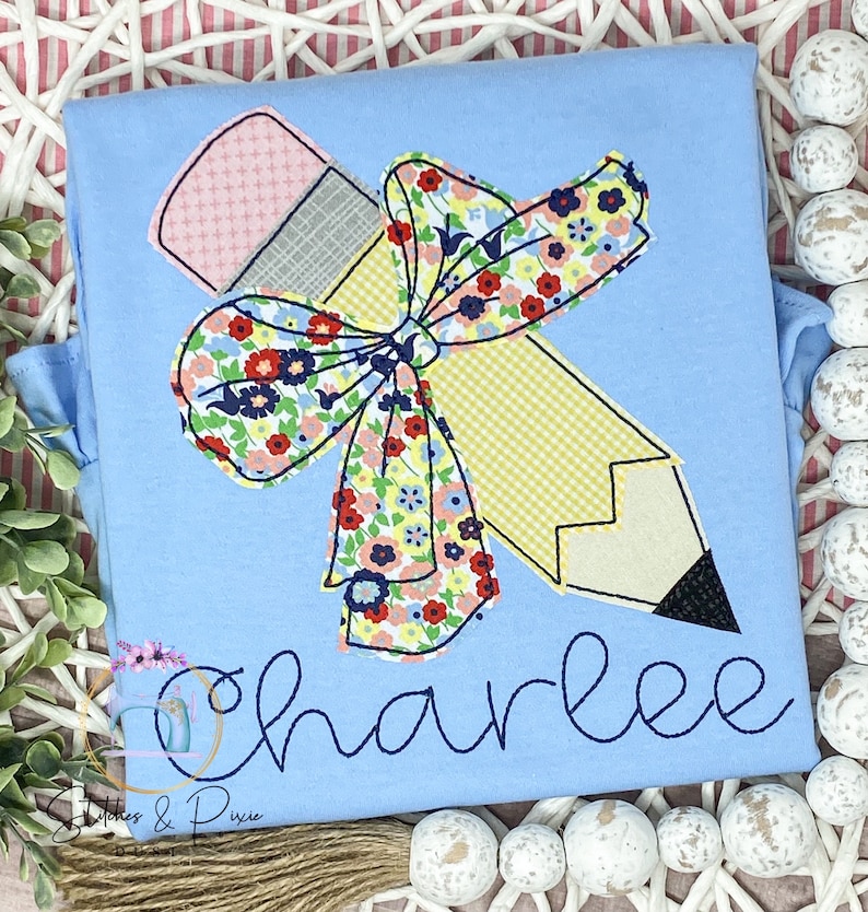 May include: A light blue t-shirt featuring a yellow pencil and a floral bow. The name "Charlee" is embroidered on the shirt.