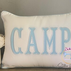 May include: White pillow with a light blue embroidered word "CAMP" on it. The pillow has a light blue border.