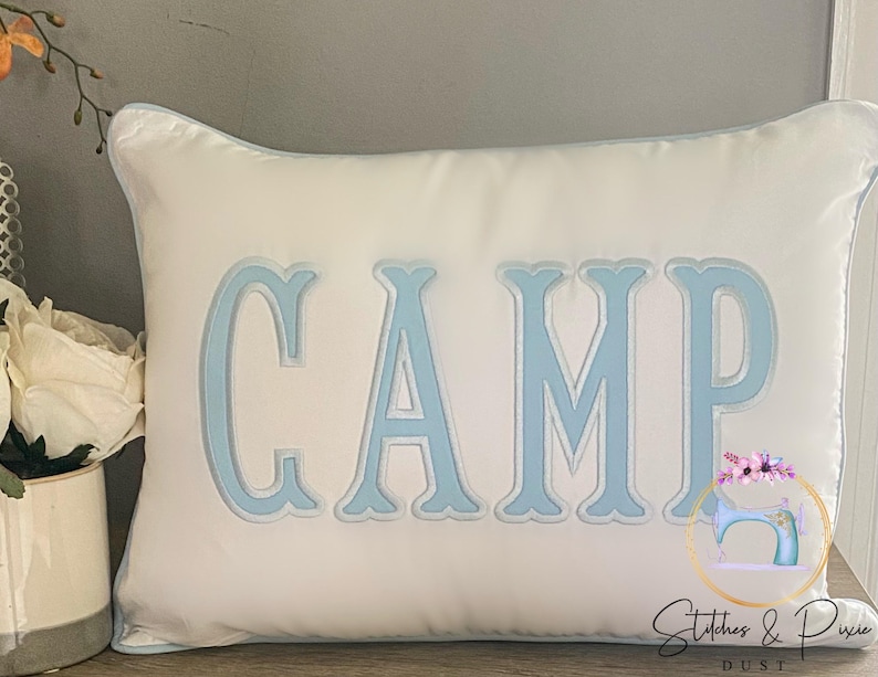 May include: White pillow with a light blue embroidered word "CAMP" on it. The pillow has a light blue border.