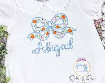 May include: White baby onesie with a blue embroidered bow with orange and pink pumpkins. The onesie has a scalloped collar and the name Abigail is embroidered below the bow.