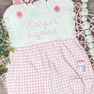 May include: Pink and white gingham baby romper with white ruffle trim and a white panel on the bodice. The panel has pink buttons and the name "Harper Lynne" embroidered in pink thread.