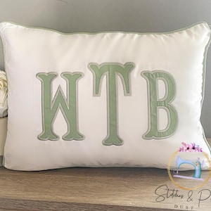 May include: White pillow with green embroidered letters that spell out "WTB".