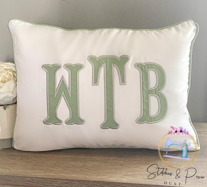 May include: White pillow with green embroidered letters that spell out "WTB".