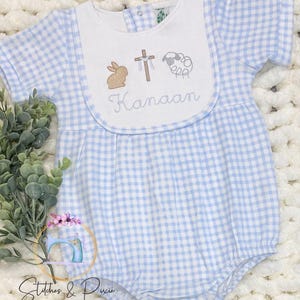 May include: Baby romper in a light blue and white gingham pattern. The white bib features embroidered images of a bunny, cross, and sheep, with the name "Kanaan" stitched below. The romper has short sleeves and a round neckline.