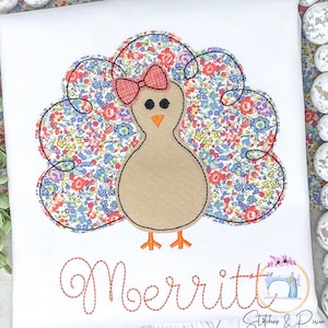 May include: A white onesie with a brown and orange turkey applique. The turkey has a floral patterned tail and a pink bow. The name "Merritt" is embroidered in orange thread below the turkey.
