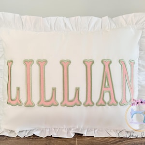 May include: A white rectangular pillow with a ruffled edge. The name "LILLIAN" is embroidered in pink letters with a green outline across the center. The pillow is on a wooden surface.