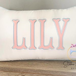 May include: White rectangular pillow with the name "LILY" embroidered in pink with a silver outline. The pillow has a light blue trim and is on a wooden surface. A logo with a sewing machine is visible.