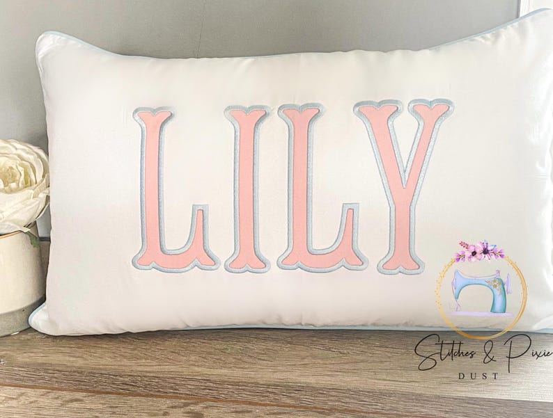 May include: White rectangular pillow with the name "LILY" embroidered in pink with a silver outline. The pillow has a light blue trim and is on a wooden surface. A logo with a sewing machine is visible.