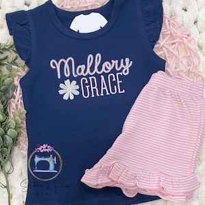 May include: Navy blue sleeveless top and pink striped shorts set. The top features the embroidered name "Mallory GRACE" with a daisy and ruffle sleeves. The shorts have a ruffled hem. The set includes the logo "Stitches & Pixie Dust".