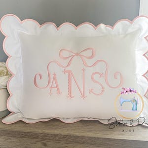 May include: A white pillow with a pink scalloped edge and pink embroidered lettering that says "ANS" with a pink bow above the letters. The pillow is on a wooden surface.