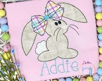 May include: A pink shirt with an embroidered brown bunny wearing a plaid bow. The bunny is sitting down and the name "Addie" is embroidered in teal below it.