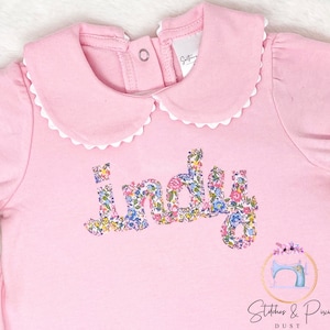 Pu&ograve; includere: Tutina rosa per neonato con colletto bianco a balze e il nome "Indy" scritto in tessuto floreale.