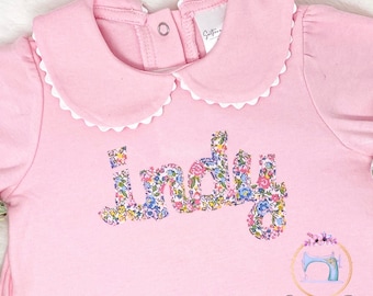 May include: Pink baby onesie with a white scalloped collar and the name "Indy" spelled out in floral fabric.