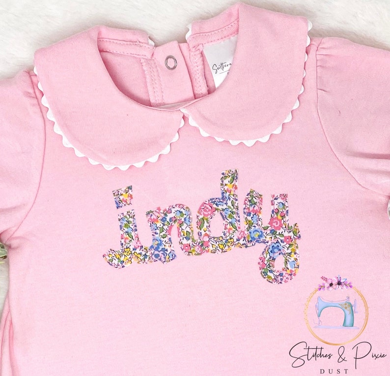Pu&ograve; includere: Tutina rosa per neonato con colletto bianco a balze e il nome "Indy" scritto in tessuto floreale.