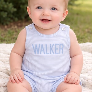 May include: A baby wearing a light blue and white striped romper with the name "WALKER" embroidered in blue. The baby is sitting on a white blanket and smiling. The romper has two small buttons on the shoulders.