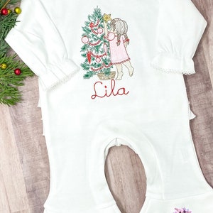May include: White long-sleeved baby onesie with a Christmas tree and a girl decorating it. The onesie has a ruffled bottom and the name "Lila" is embroidered on the front.