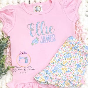 May include: Pink ruffle-sleeved top and floral shorts set. The top features the name "Ellie James" in blue script, a floral design, and the logo "Stitches & Pixie Dust". The shorts have a blue, yellow, and white floral pattern.