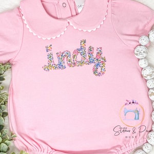 Pu&ograve; includere: Tutina rosa per neonato con un colletto bianco a balze e il nome "Indy" scritto in lettere floreali. La tutina ha un fondo a balze e un piccolo logo di macchina da cucire in basso a destra.