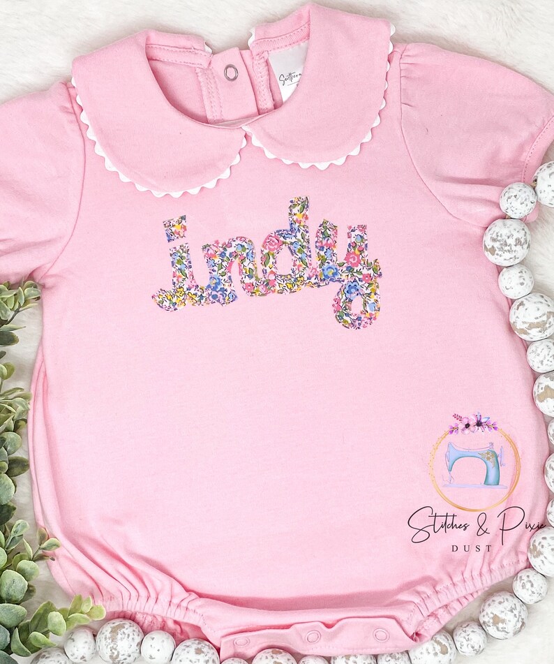 Pu&ograve; includere: Tutina rosa per neonato con un colletto bianco a balze e il nome "Indy" scritto in lettere floreali. La tutina ha un fondo a balze e un piccolo logo di macchina da cucire in basso a destra.