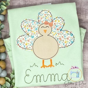 May include: A light green shirt featuring a floral patterned turkey applique. The turkey has a pink bow and the name "Emma" is embroidered beneath it.