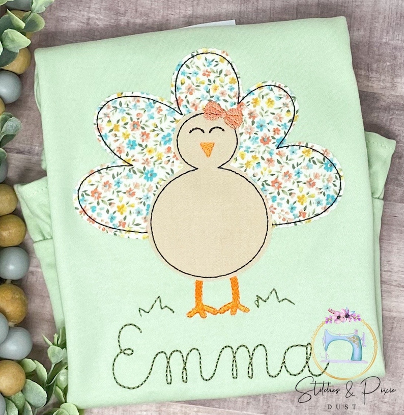 May include: A light green shirt featuring a floral patterned turkey applique. The turkey has a pink bow and the name "Emma" is embroidered beneath it.