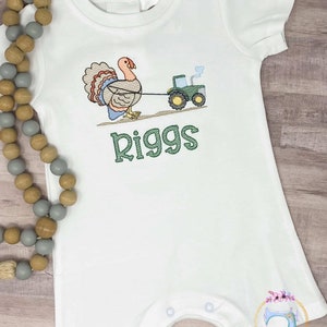 May include: White baby onesie with a green tractor pulling a turkey. The onesie has the name "Riggs" embroidered in green.