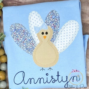 May include: A light blue t-shirt with a brown and beige applique of a turkey with a blue bow. The turkey has a floral patterned tail and white wings with blue polka dots. The name "Amisty" is embroidered in blue cursive lettering below the turkey.