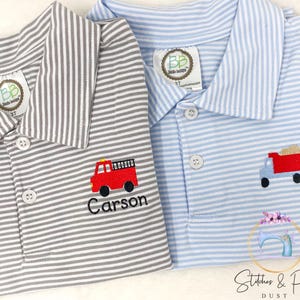May include: Two striped polo shirts, one gray and one blue, with embroidered images of a fire truck and a dump truck. The gray shirt has the name "Carson" embroidered below the fire truck.