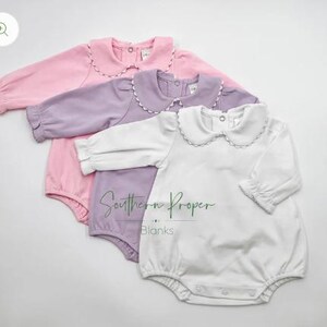 Pu&ograve; includere: Tre body per neonati con colletti a volant. I body sono rosa, lavanda e bianco. I body hanno maniche lunghe e chiusure a pressione.