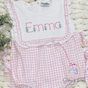 May include: A white and pink gingham baby romper with a white bib. The bib features the name "Emma" embroidered in pink with blue flower accents. The romper has ruffled edges and tie straps.