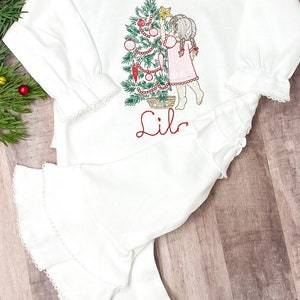 May include: White long-sleeved shirt with a ruffled bottom and a Christmas tree design featuring a girl decorating the tree. The shirt is personalized with the name "Lila".