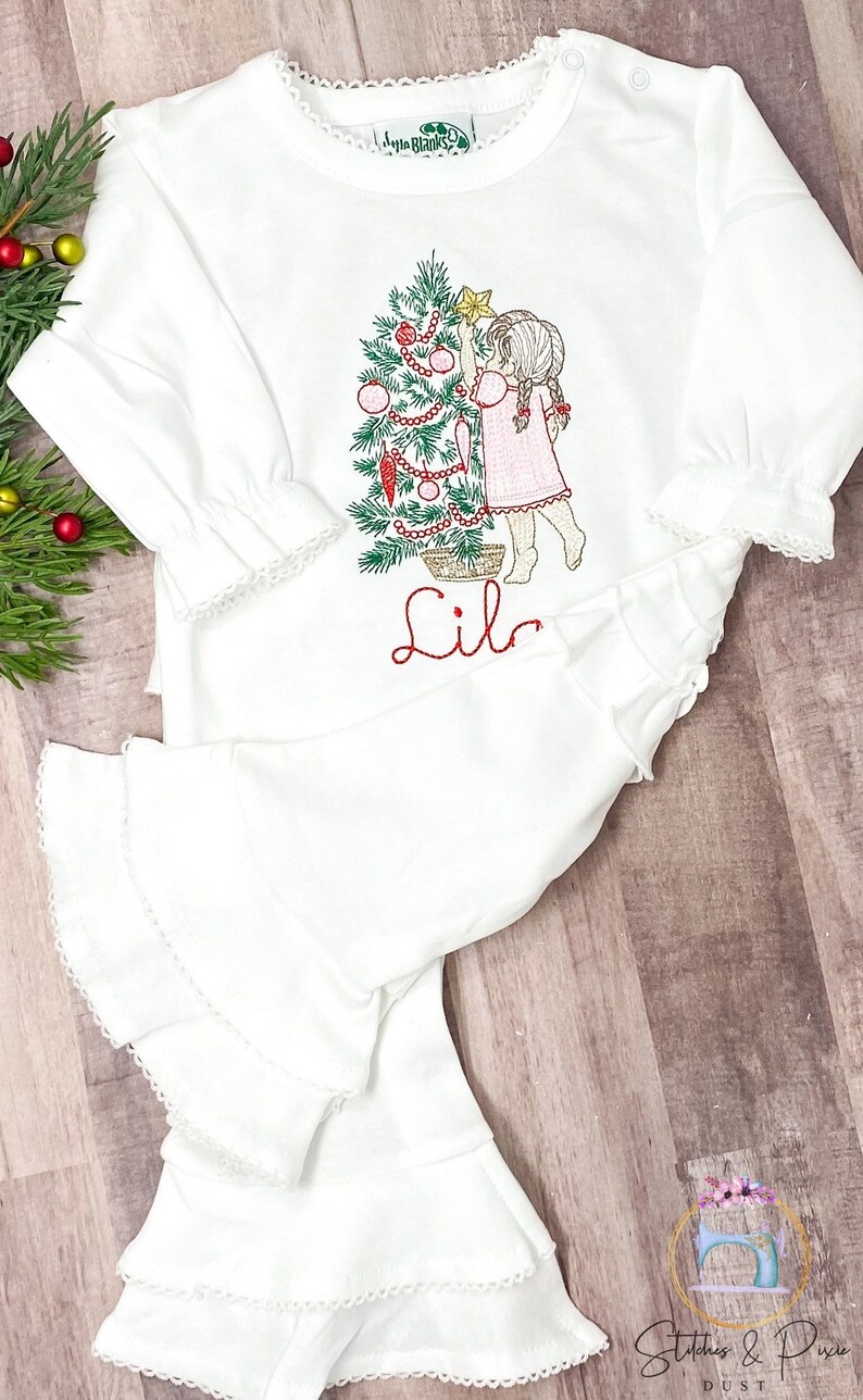 May include: White long-sleeved shirt with a ruffled bottom and a Christmas tree design featuring a girl decorating the tree. The shirt is personalized with the name "Lila".