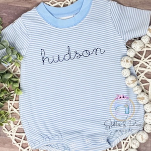 May include: A light blue and white striped baby onesie with the name "hudson" embroidered in black on the front. The onesie has a blue trim around the neckline.