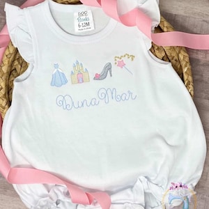 May include: White baby onesie with a blue embroidered design of a princess, castle, shoe, and wand. The onesie has the name "Luna Noar" embroidered in blue below the design. The onesie is size 6-12M.