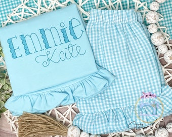 May include: A light blue baby outfit with a ruffled hem. The top has the name "Emnie Kate" embroidered on it. The bottom is a gingham print with a ruffled hem.