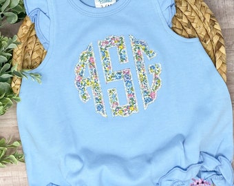 May include: A light blue baby onesie with ruffled sleeves and a floral monogram on the front. The onesie has a snap closure at the bottom.