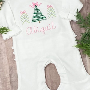 May include: White baby onesie with a green embroidered Christmas tree and pink bows. The onesie has a ruffled bottom and the name "Abigail" is embroidered below the tree.