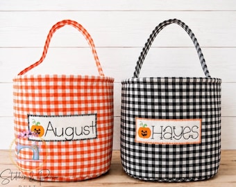 May include: Two cylindrical fabric baskets with handles, one in orange and white gingham and one in black and white gingham. Each features a rectangular white patch with an embroidered orange jack-o'-lantern and the names August and Hayes.