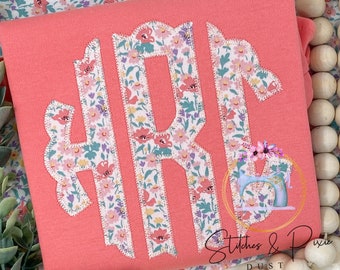 May include: A coral pink t-shirt with a white monogram in a floral print. The monogram is a large "R" with a scalloped edge. The t-shirt is made of a soft, comfortable fabric. The text "Stitches & Pixie Dust" is visible in the bottom right corner of the image.