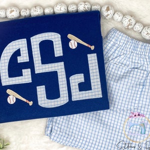 May include: A navy blue t-shirt with a white monogram and baseball graphic. The monogram is in a gingham pattern. The t-shirt is paired with blue and white gingham shorts.