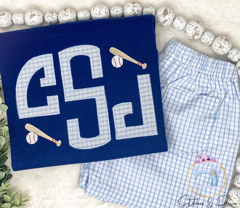 May include: A navy blue t-shirt with a white monogram and baseball graphic. The monogram is in a gingham pattern. The t-shirt is paired with blue and white gingham shorts.