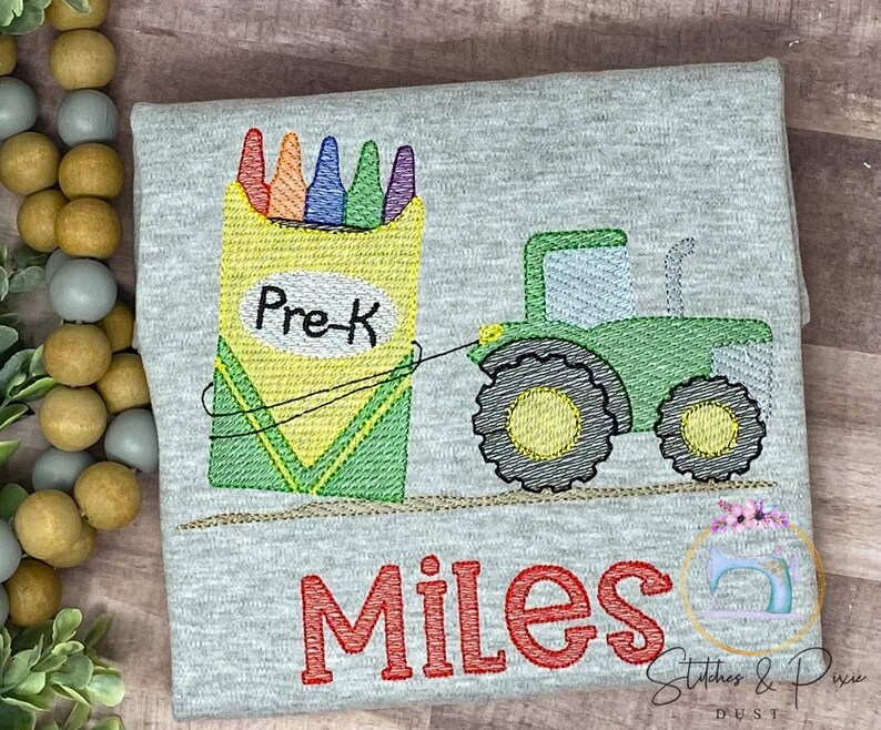 May include: A gray t-shirt with an embroidered design of a green tractor pulling a box of crayons. The box of crayons has the words "Pre-K" on it. The name "Miles" is embroidered below the tractor.