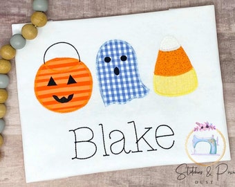 May include: White t-shirt with an orange pumpkin, blue ghost, and yellow candy corn applique. The name "Blake" is embroidered in black below the appliques.