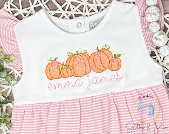 Pode incluir: Vestido branco sem mangas com listras rosa e brancas. O vestido tem um corpete branco com abóboras bordadas e os nomes "emma james" em fio rosa.
