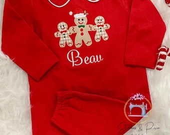 May include: Red baby romper with long sleeves and a white-trimmed collar. The romper features an embroidered design of three gingerbread people and the name "Beau" in cursive. A matching red diaper cover is included.