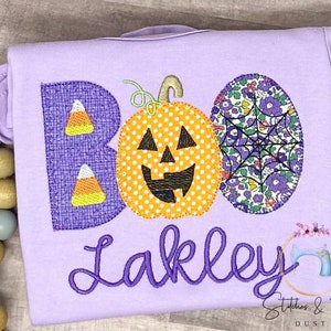 May include: Purple T-shirt with the word "BOO" embroidered in purple featuring a pumpkin, candy corn, and spiderweb designs. The name "Lakley" is embroidered in purple below the word "BOO".