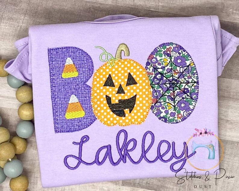 May include: Purple T-shirt with the word "BOO" embroidered in purple featuring a pumpkin, candy corn, and spiderweb designs. The name "Lakley" is embroidered in purple below the word "BOO".