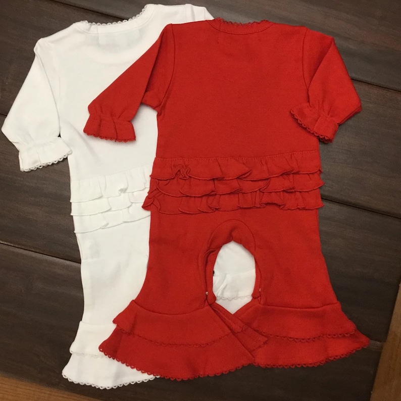 May include: Two baby onesies, one white and one red, with ruffled trim around the neck, sleeves, and bottom. Both onesies have a snap closure at the crotch.