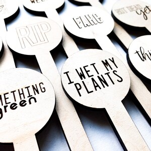 May include: A set of wooden plant markers with humorous and irreverent phrases, including "RIP", "I Wet My Plants", "Something Green", "Thinsty B*tch", and "Shot Dead Yet".