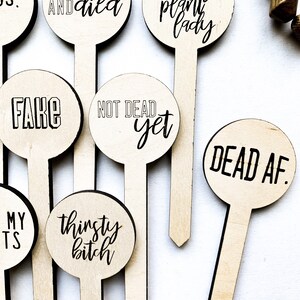 May include: Set of 8 wooden plant stakes with funny sayings. The stakes are round and have black text on a natural wood background. The sayings include "Tried and Died", "Not a Crazy Plant Lady", "Fake", "Not Dead Yet", "Dead AF", "Thirsty Bitch", "My T's" and "DS."